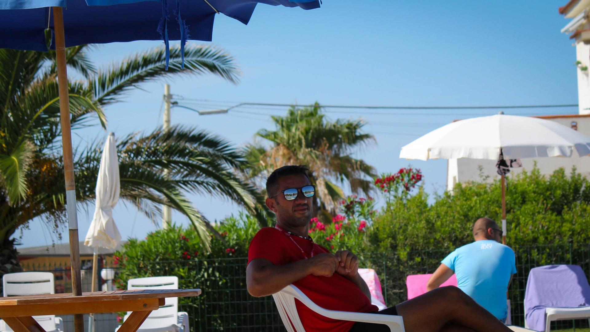 Uomo in spiaggia sotto l'ombrellone, indossa occhiali da sole e maglietta rossa.