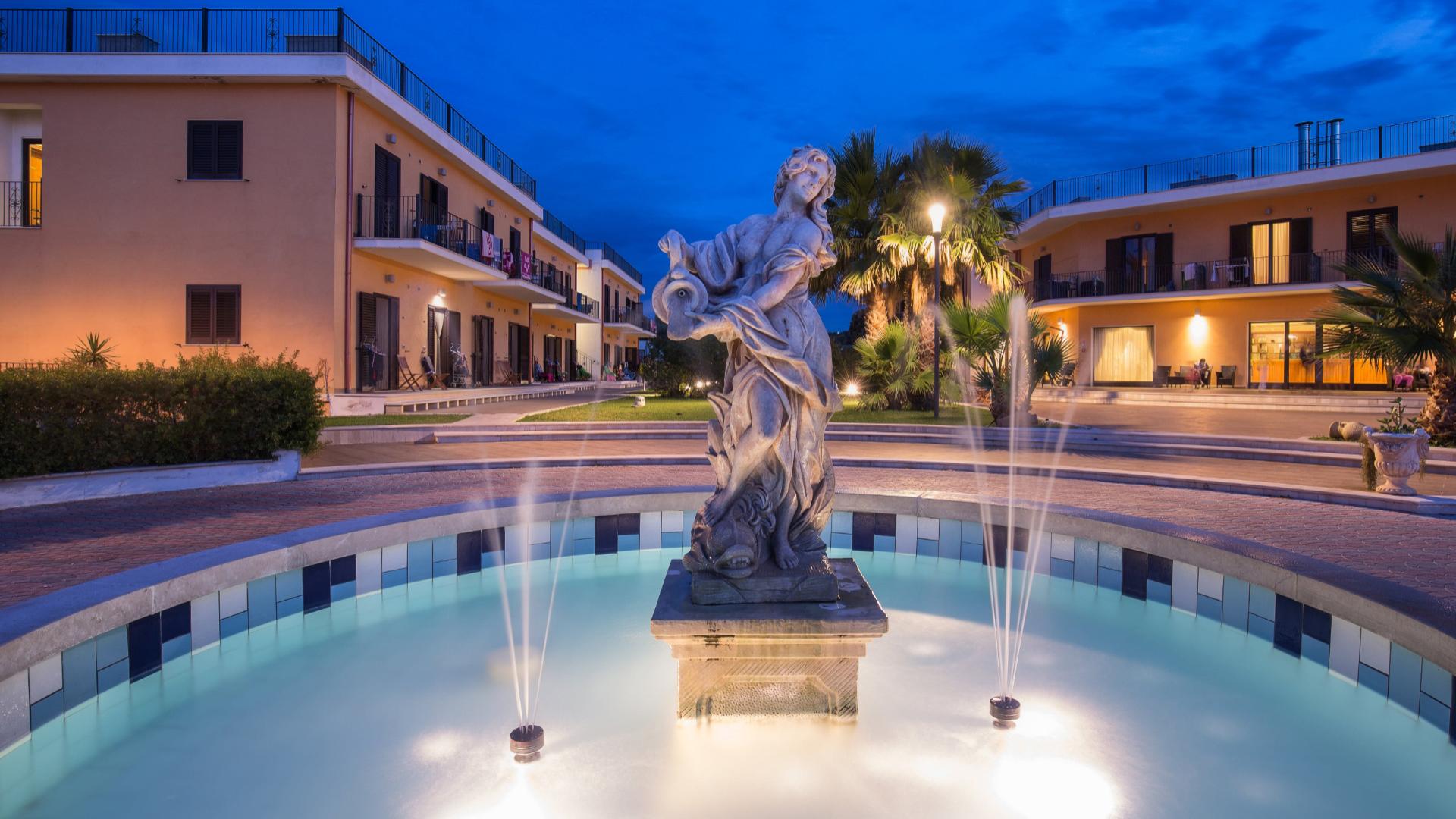 Fontana con statua al centro di un cortile illuminato di sera.