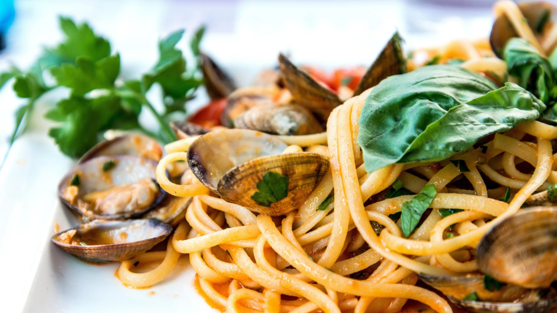Spaghetti alle vongole con pomodoro, basilico e prezzemolo su piatto bianco.