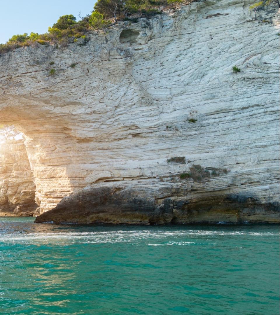 Scogliera bianca con arco naturale e mare turchese, illuminata dalla luce del sole.