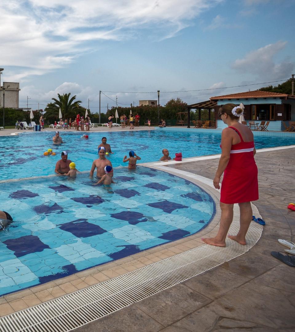 Persone che giocano in piscina, sorvegliate da una donna in abito rosso.