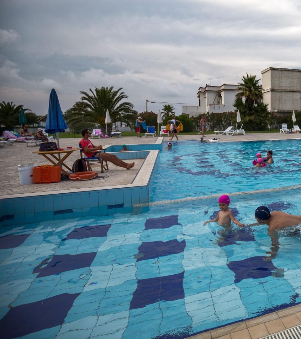 Piscina all'aperto con bagnino, bambini che nuotano e persone che si rilassano.