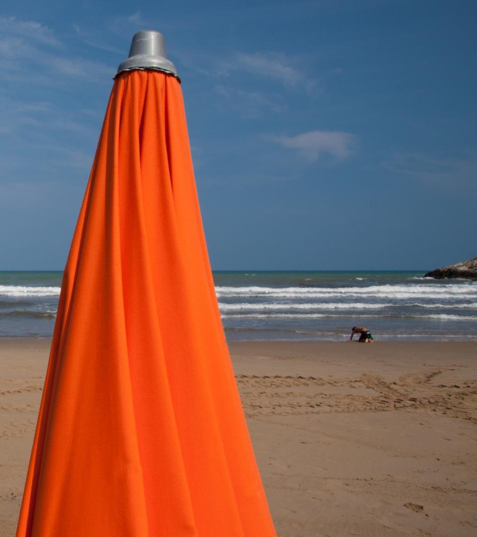 Ombrellone arancione chiuso su una spiaggia con mare e cielo sereno sullo sfondo.