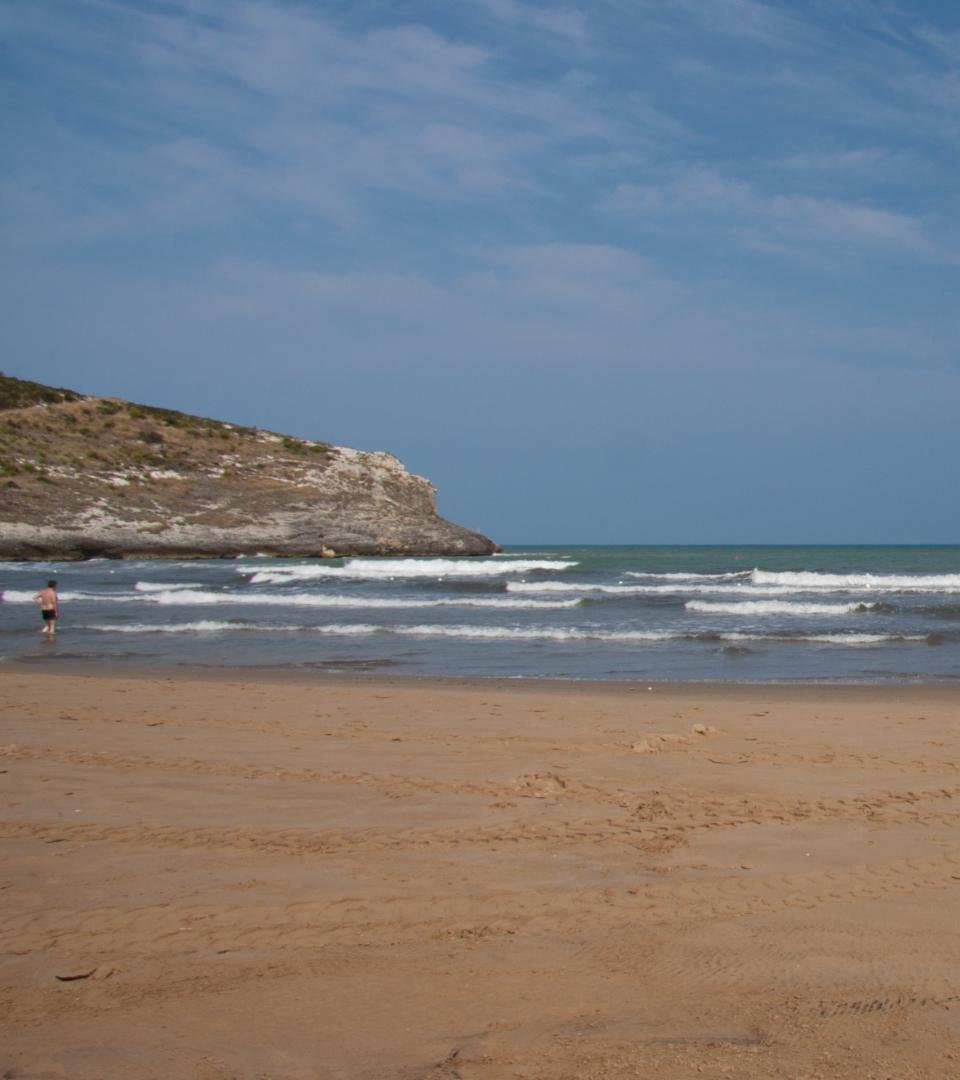 Spiaggia sabbiosa con onde e una scogliera sullo sfondo in una giornata soleggiata.